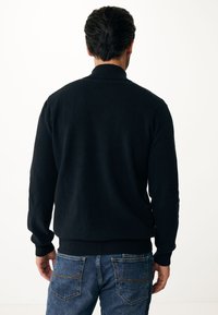 Svart stickad tröja med hög krage, ribbade ärmslut och nederkant. Bärs över blå denimjeans. Mjuk textur, enkel design. Bakvy.