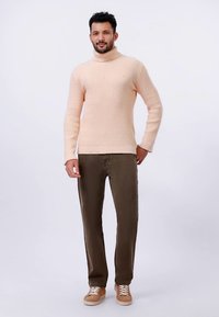 Lys rosa strikket genser med høy hals og ribbestrikkede mansjetter, kombinert med olivengrønne bukser og beige sneakers, stående mot en ensfarget bakgrunn.