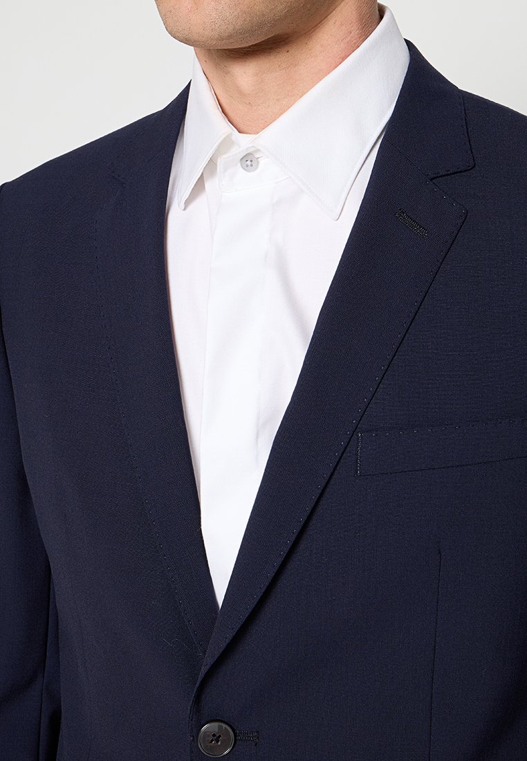 Mann in einem dunkelblauen Blazer über einem weißen Hemd mit spitzem Kragen, das oben geknöpft ist.