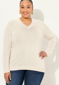 V-Ausschnitt Pullover mit langen Ärmeln in hellem Beige, feinem Strickmuster, verlängerter Passform und gerippten Saumdetails. Kombiniert mit blauen Jeans.