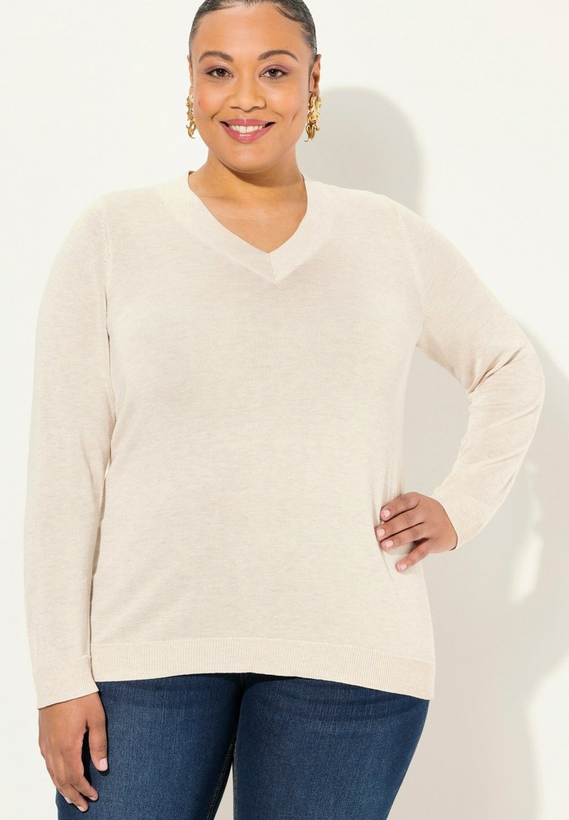 V-Ausschnitt Pullover mit langen Ärmeln in hellem Beige, feinem Strickmuster, verlängerter Passform und gerippten Saumdetails. Kombiniert mit blauen Jeans.