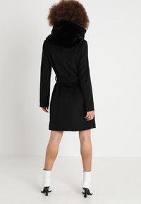Abrigo negro con un gran cuello de terciopelo negro, cintura ceñida con cinturón, forma ajustada y bolsillos laterales, combinado con brillantes botas de tobillo blancas.