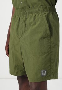 Shorts vert olive avec une ceinture élastique, des poches latérales et un patch logo GAP en forme rectangulaire. Tissu texturé et léger.