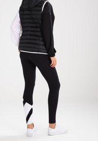 Černé legíny s bílým a černým geometrickým vzorem na spodní části nohy, v kombinaci s černou prošívanou vestou a bílými teniskami.