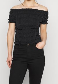 Svart off-shoulder topp med en texturerad krosserdesign, som har korta ärmar och en figurnära silhuett. I kombination med svarta jeans.