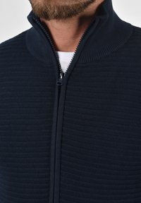 Marineblå lynlåstrøje med ribbet tekstur, høj krave og kontrasterende lynlås. Stoffet ser blødt og let strækbart ud.