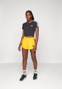 Μαύρη κοντή αθλητική μπλούζα με λευκές ρίγες στους ώμους, σε συνδυασμό με κίτρινα σορτς τρεξίματος με μαύρες λεπτομέρειες και λογότυπο. Μαύρα αθλητικά παπούτσια.