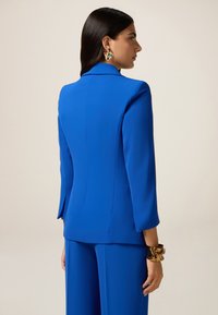 Royal blue blazer made of smooth fabric, featuring a tailored fit, notched collar, and three-quarter sleeves. Gold statement earrings and bracelet.