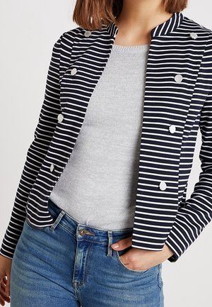 Femme portant un blazer croisé à rayures bleu marine et blanc sur un haut gris clair et un jean bleu, une main dans la poche.