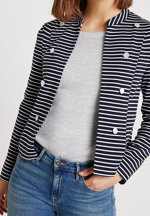 Femme portant un blazer croisé à rayures bleu marine et blanc sur un haut gris clair et un jean bleu, une main dans la poche.