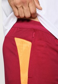 Dunkelrote Sportshorts mit orange gemustertem seitlichem Einsatz, strukturiertem Stoff, mit verdecktem Reißverschlusstasche in der Nähe des Taillenbunds.