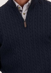 Marineblauwe gebreide sweater met een textuur kabelontwerp, voorzien van een kwart-rits nek en een leren ritsing met een bruine kleur. Eronder is een gemusterd shirt gedeeltelijk zichtbaar.