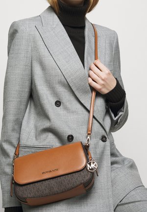 Femme portant un blazer gris croisé et un col roulé noir, tenant un sac bandoulière Michael Kors marron à motifs foncés.