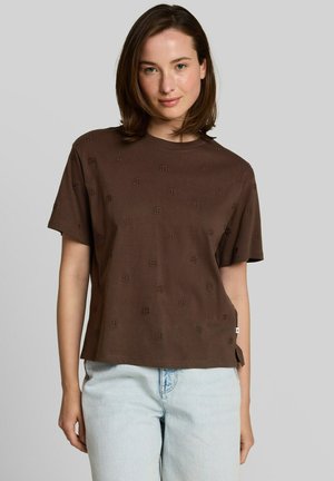 Jonge vrouw met schouderlang kastanjebruin haar draagt een bruin shirt met patroon en korte mouwen en lichtblauwe spijkerbroek, staand tegen een effen achtergrond.