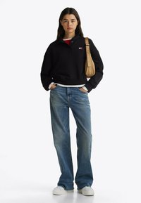 Zwarte cropped sweatshirt met kraag, knoopdetails en logo. Gecombineerd met blauwe wijde jeans en witte Sneakers. Tan tas met logo-accenten.