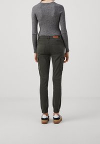 Pantalones cargo en verde oscuro con puños elásticos, que presentan bolsillos en los lados y una etiqueta en la parte posterior. Combinados con una camiseta ajustada de canalé gris y zapatos casuales.