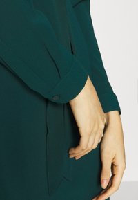 Ciemnozielona bluzka z miękkiego, teksturowanego materiału. Posiada zapięcie na guzik przy mankietach oraz detal w postaci panelu z boku, co podkreśla luźny krój.