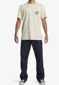 Krämfärgad bomullströja med grön logotyp på vänster bröst, tillsammans med mörkblå denimjeans och svartvita sneakers.