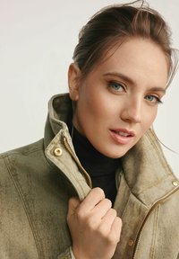 Beżowa dżinsowa kurtka z wysokim kołnierzem, złotymi zamkami i teksturowanym materiałem. Czarny golf pod spodem. Czyste, minimalistyczne tło.