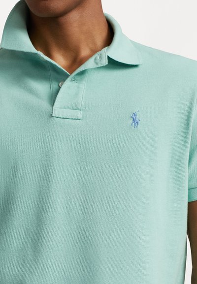 Polo majice v mint zeleni barvi, narejene iz teksturirane tkanine, z klasikim ovratnikom, dvema gumboma in majhnim vezenim logotipom na prsih.