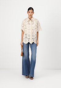 Blusa in pizzo bianco sporco con motivo floreale, maniche corte e orlo scallop; abbinata a jeans a zampa d'elefante blu e sandali neri.