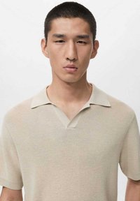 Beżowa koszulka polo z krótkim rękawem o teksturowanym splocie, z kołnierzykiem z wycięciem i delikatnym prążkowaniem. Prosty fason, odpowiedni na co dzień.