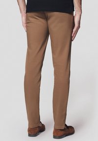 Brązowe chinosy wykonane z gładkiego materiału, w fasonie slim fit, z kieszeniami z tyłu i kontrastowymi brązowymi butami. Neutralne tło.