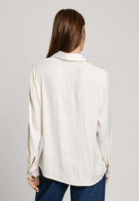 Blusa a maniche lunghe di colore crema con colletto, dalla texture liscia e leggermente lucida. Presenta un orlo curvo e polsini abbottonati, indossata con jeans.