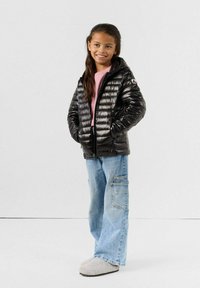 Chaqueta de plumas negra con textura acolchada, capucha, un logo en la manga, combinada con pantalones cargo de pierna ancha en azul claro y zapatillas grises.