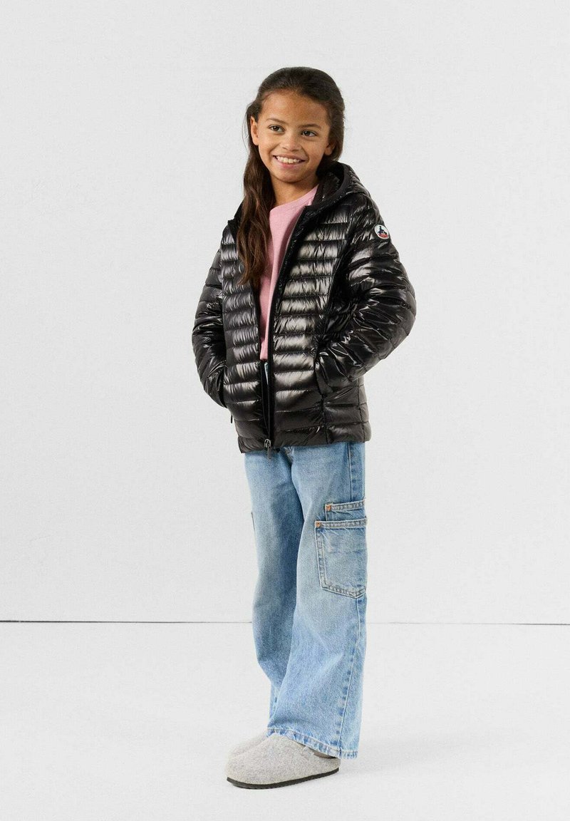 Chaqueta de plumas negra con textura acolchada, capucha, un logo en la manga, combinada con pantalones cargo de pierna ancha en azul claro y zapatillas grises.