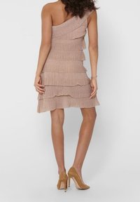 Kvinna i en ärmlös, enaxlad beige lagerklänning och beige högklackade skor, som står med ryggen vänd mot betraktaren.