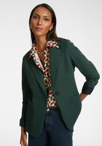 Donna che indossa un blazer verde scuro sopra una blusa a fantasia e jeans scuri, in posa con una mano nella tasca contro uno sfondo bianco.