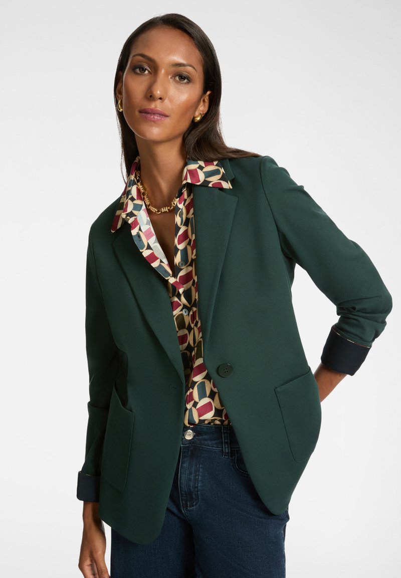 Donna che indossa un blazer verde scuro sopra una blusa a fantasia e jeans scuri, in posa con una mano nella tasca contro uno sfondo bianco.