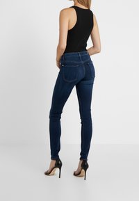 Jeans ajustados de color azul oscuro con cintura alta, confeccionados en denim. Presentan dos bolsillos traseros y detalles de costura sutiles. El calzado incluye tacones negros.