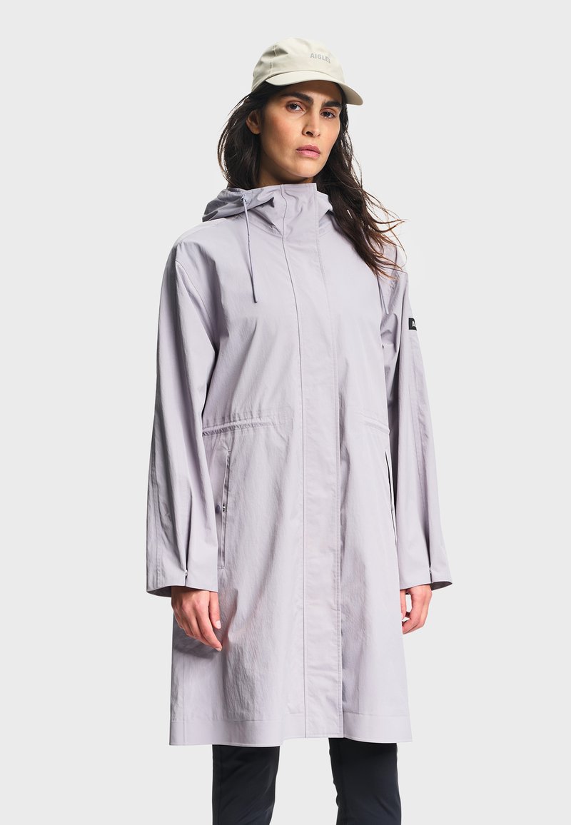 Femme portant un imperméable à capuche gris clair et une casquette beige, debout devant un fond blanc uni.