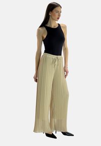 Zwarte mouwloze top, geplooide beige wijde broek met een trekkoord in de taille en zwarte puntige hakken. Glad stof met verticale textuur.