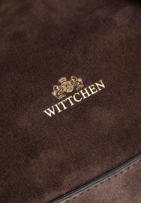 Materiale in suede marrone con una superficie liscia. Presenta un logo dorato "WITTCHEN" e uno stemma, offrendo un dettaglio di alta qualità.