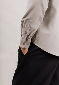 Lys grå kordfløyelskjorte med rundet kant, knapping ved ermer og jevn tekstur, kombinert med svarte bukser. Hånd i lomma.