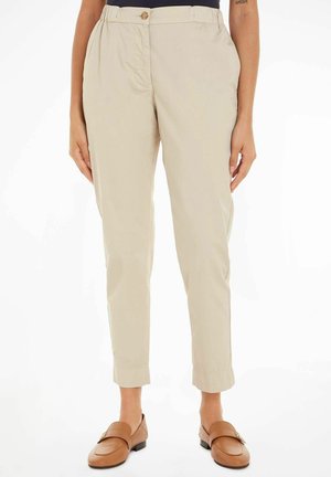 Pantaloni beige a gamba affusolata con chiusura a bottone, tasche laterali e una texture liscia. Indossati con mocassini marrone chiaro.