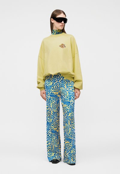 Femme portant un sweat-shirt jaune clair surdimensionné, un pantalon large à motifs bleus et jaunes, des lunettes de soleil noires et des chaussures noires, debout devant un fond blanc.