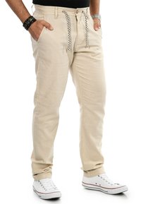 Beige Hosen mit schmalem Schnitt, ausgestattet mit einem gewebten Kordelzug in Schwarz-Weiß-Muster, seitlichen Taschen und umschlägigen Säumen. Kombiniert mit weißen Turnschuhen.