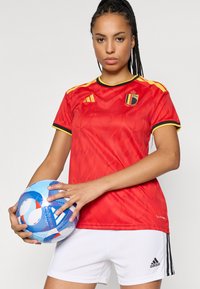 Κόκκινη ποδοσφαιρική φανέλα με κίτρινες λεπτομέρειες, που διαθέτει έμβλημα και μαύρες ρίγες. Συνδυάζεται με λευκά σορτς και κρατάει μια μπλε ποδοσφαιρική μπάλα με σχέδιο.