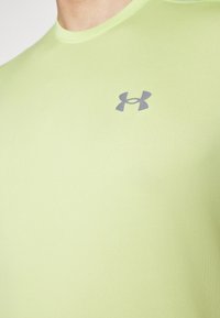 Jasnozielona dopasowana koszulka z miękkiego materiału, o gładkiej fakturze i szarym logo Under Armour na górnej lewej części klatki piersiowej.