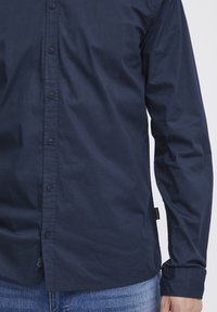 Camicia in cotone blu navy con bottoni, caratterizzata da maniche lunghe, colletto classico e orlo dritto con una piccola etichetta del marchio sul lato.
