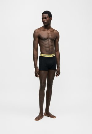 Slim muscular black man standing barefoot wearing black boxer briefs with yellow waistband that reads "SWEDEN BJÖRN".