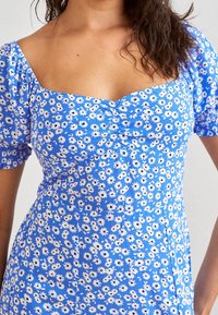 Blaues Blumenmusterkleid mit kurzen Puffärmeln, das über einen quadratischen Ausschnitt und geraffte Details verfügt. Weiße Gänseblümchenmuster auf der gesamten Fläche. Texturierter Stoff.