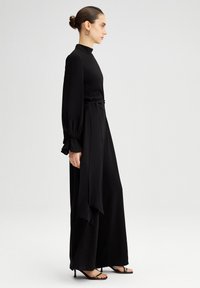 Salopetă neagră cu mâneci lungi, guler înalt, pantaloni largi și un detaliu legat la talie. Materialul pare neted, cu o textură fluidă.