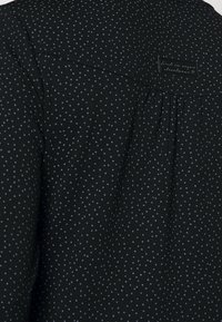 Svart tygblus med ett mönster av små ljusblå prickar, med ett plisserat detaljer bak och en varumärkesetikett sydd vid kragen.