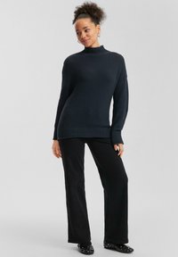 Marineblauwe geribbelde trui met hoge hals, gecombineerd met zwarte wijde broek en zwarte schoenen met zilveren studs.