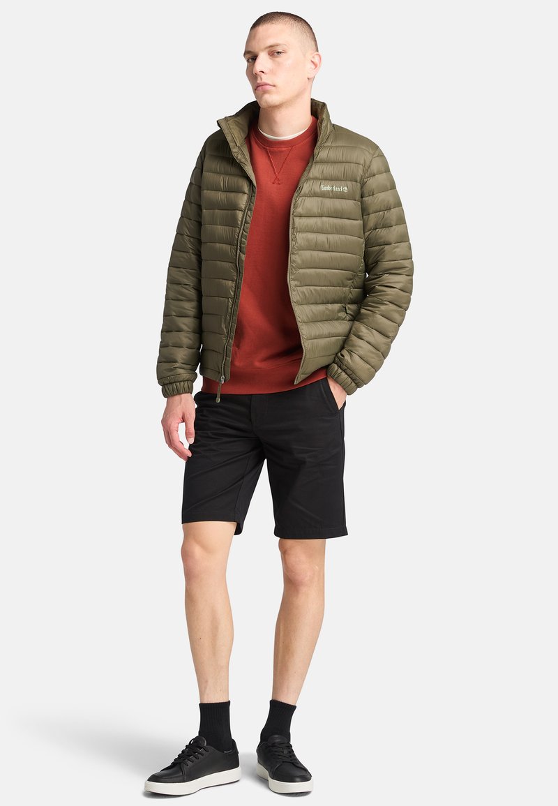 Oliwkowa kurtka puchowa z zamkiem, noszona na czerwonym swetrze, w zestawie z czarnymi szortami i czarnymi trampkami z białą podeszwą.